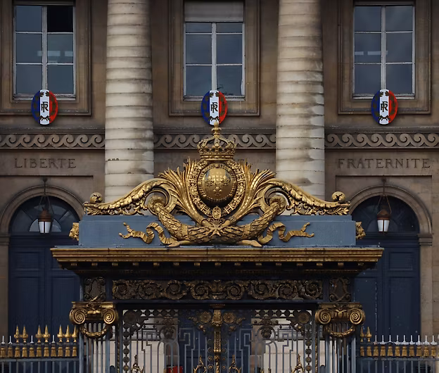 Facade of an official French building with stone columns, two windows displaying tricolor emblems, inscriptions ‘LIBERTÉ’ and ‘FRATERNITÉ’ engraved in the stone, and an ornate golden gate featuring a central crest topped with a crown.